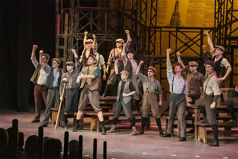 Musical theater cast performing on stage in period costumes with raised fists during a high-energy ensemble number