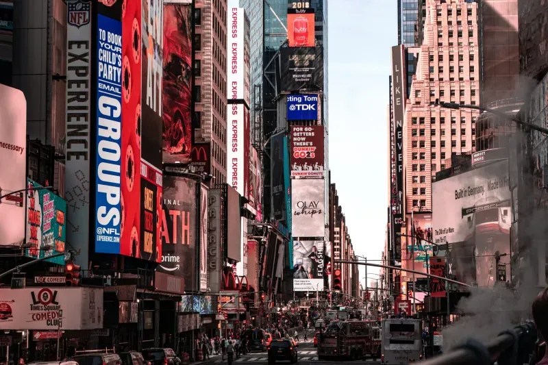 Wide-angle view of Times Square in New York City featuring towering skyscrapers covered in bright digital billboards, including advertisements for NFL Experience, Dunkin', and Olay, with city traffic and steam in the foreground