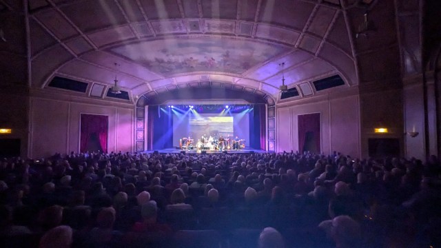 Historic theater with ornate ceiling; a band performs on a lit stage to a large audience bathed in purple light.